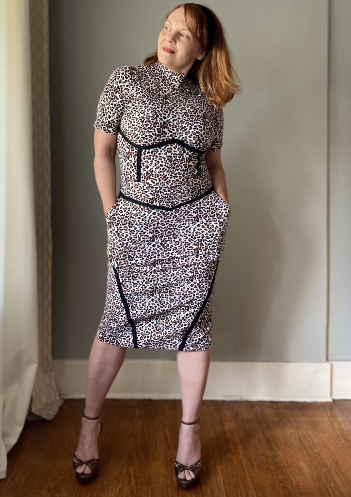 A mid-50s white woman with red hair stands in front of a grey wall looking to the left. She wears a handmade animal print top and skirt with black seam detailing.