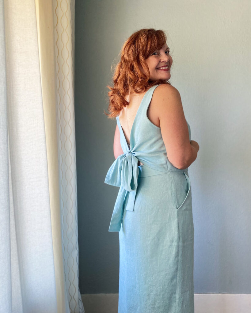 A mid-50s white woman with red hair stands facing a grey wall wearing a handmade light blue sundress.