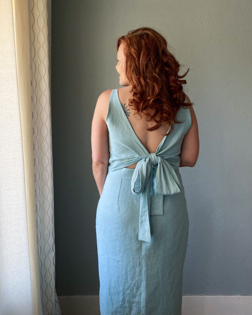 A mid-50s white woman with red hair stands facing a grey wall wearing a handmade light blue sundress.