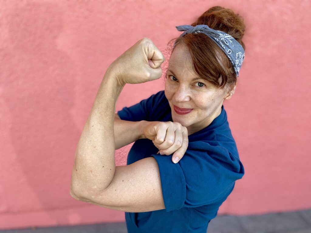 A waist-up shot of a white woman with red hair wearing a teal top against a pink wall. She makes a fist in the the style of Rosie the Riveter.