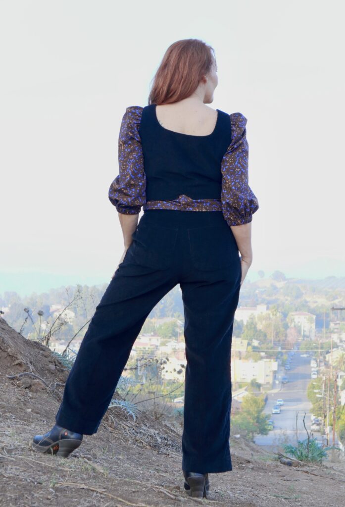 Back view of red-haired woman wearing black corduroy vest and pants with poufy-sleeved purple printed blouse.