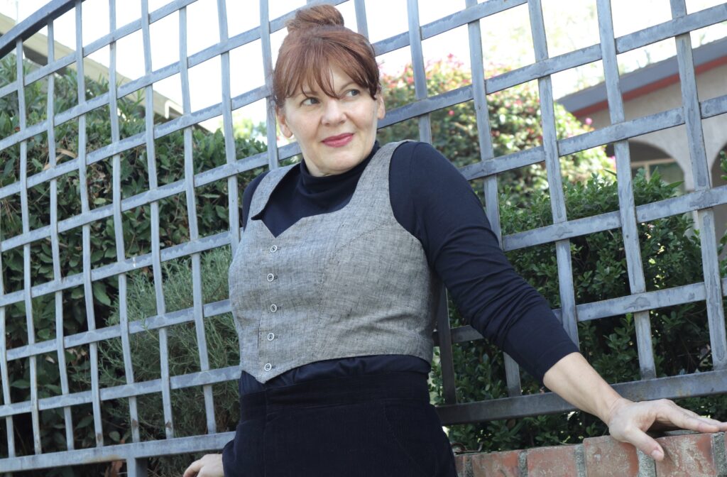 A red-haired woman leans against a metal fence wearing a grey vest and black top.