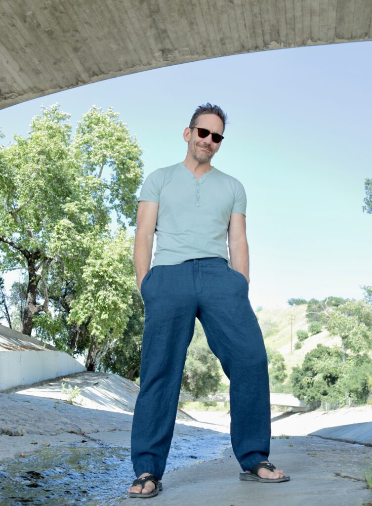 A white man wearing sunglasses, light blue shirt, and dark blue pants stands in a dry riverbed under an overpass. There are trees in the background.
