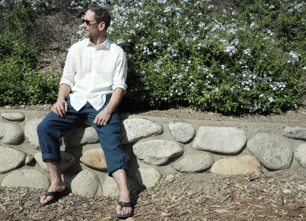 A white man wearing sunglasses, white shirt, and dark blue pants sits on a stone wall in front of a green bush with white flowers.