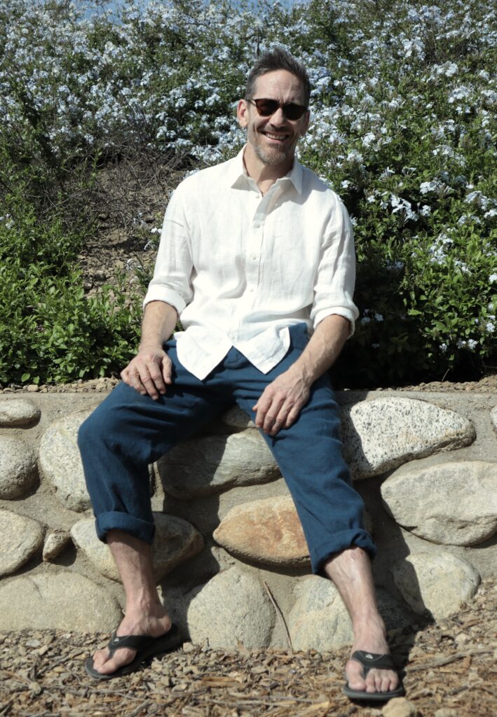 A white man wearing dark blue pants and a white shirt sits on a stone wall.