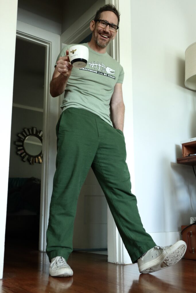 A white man wearing green pants, light green t-shirt, white shoes, and glasses holds up a coffee mug as he leans against a white door frame.