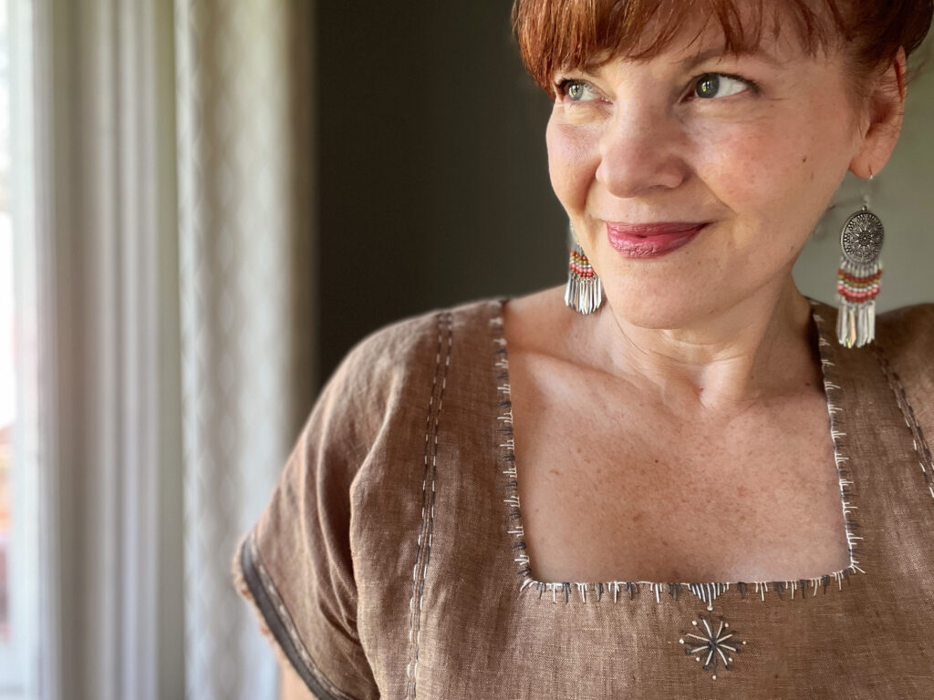 Close up of a white woman with red hair wearing a brown embroidered linen top and large beaded earrings