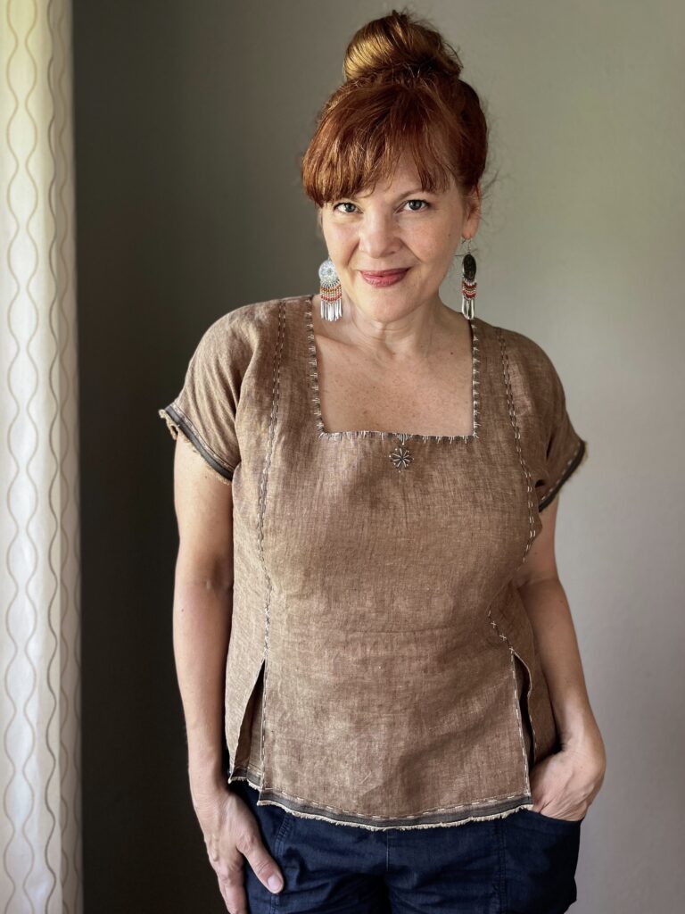 A white woman with red hair wears a brown embroidered linen top and large beaded earrings