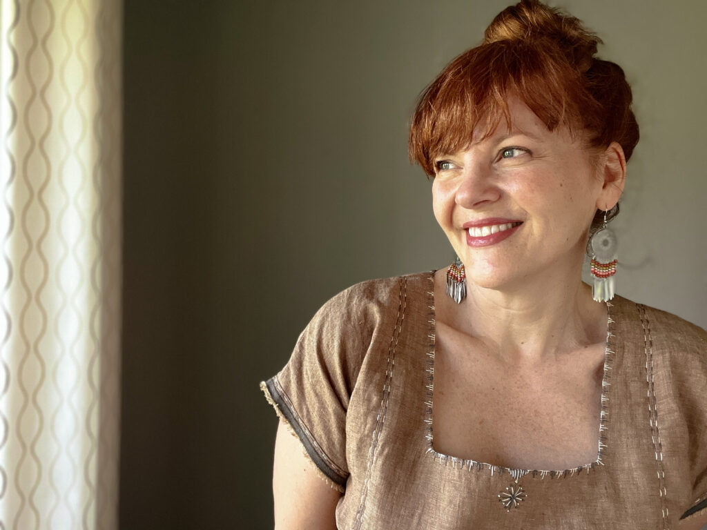 Close up of a white woman with red hair smiling  and wearing a brown embroidered top and large beaded earrings