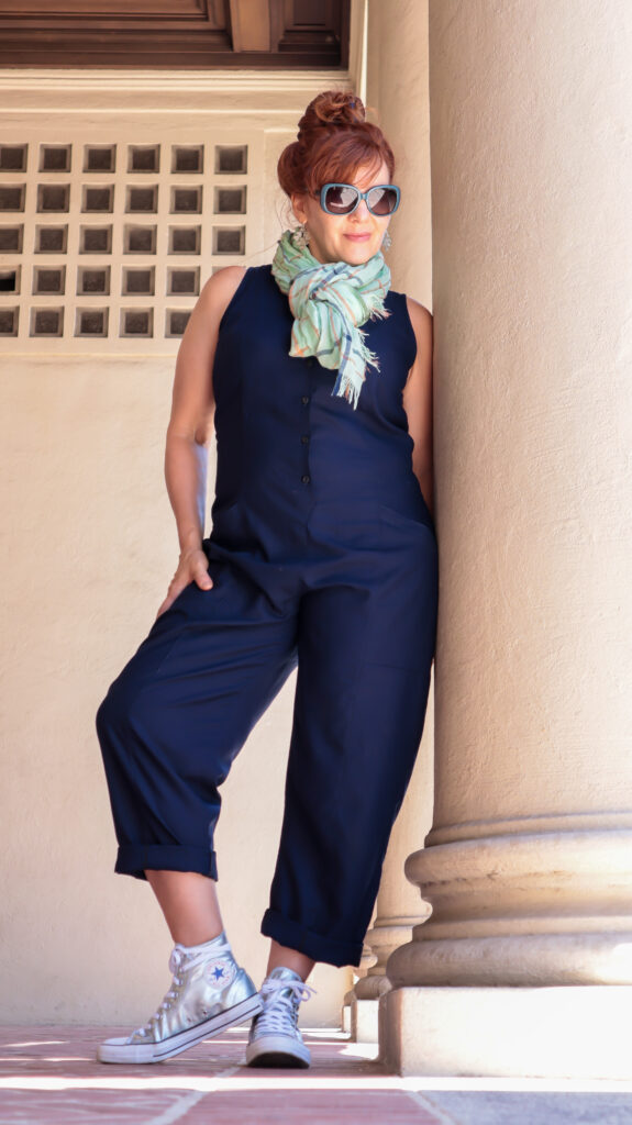 A woman with red hair leans against a stone column wearing a navy blue jumpsuit, sunglasses, mint green scarf, and silver sneakers.