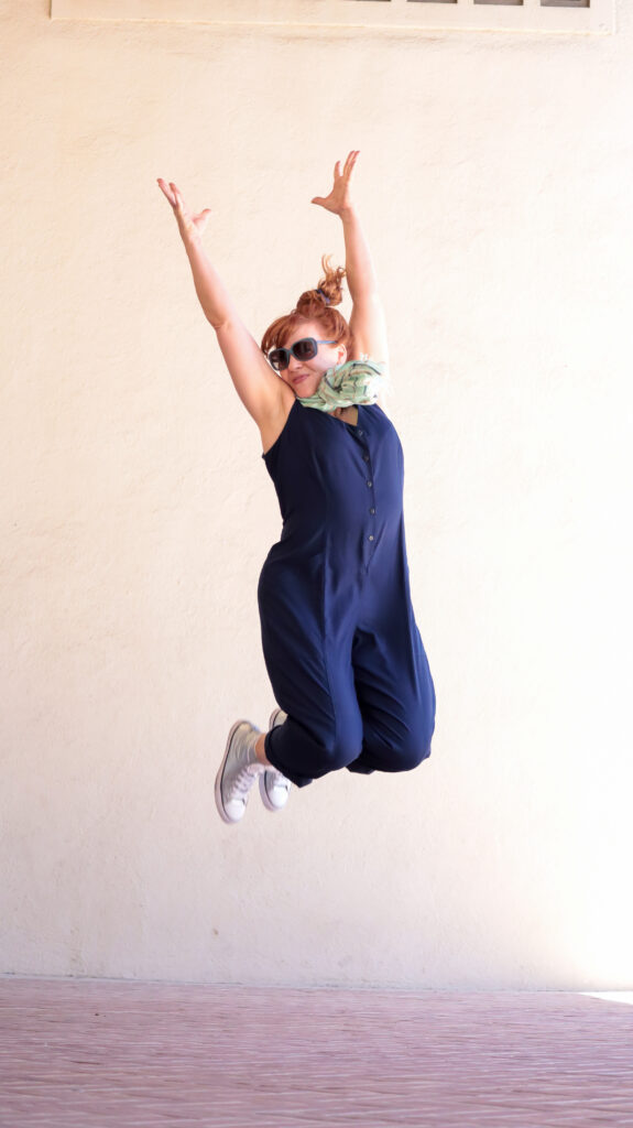 A woman with red hair jumps in the air wearing a navy blue jumpsuit, sunglasses, mint green scarf, and silver sneakers.
