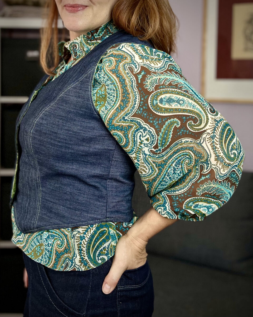 Side view of a white woman with read hair wearing a denim vest over a green and brown paisley blouse