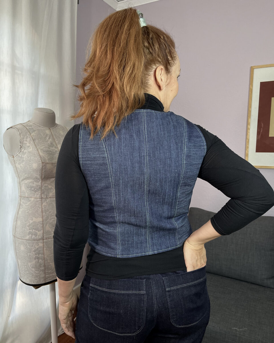 Back view of a white woman with red hair wearing a denim vest over a black turtleneck.