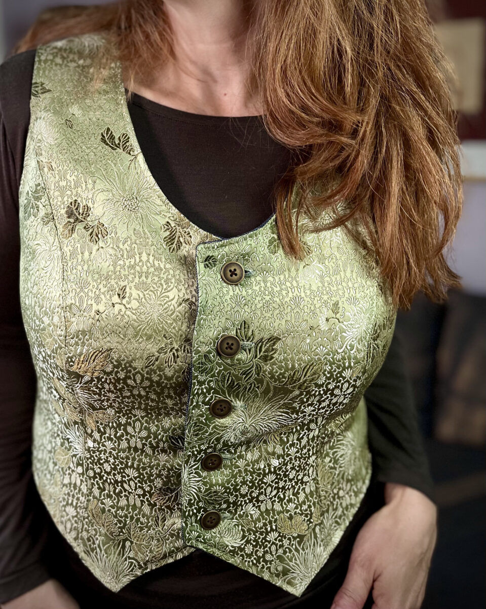 Close up of a white woman with red hair wearing a sage green silk brocade vest over an olive long sleeve t-shirt.