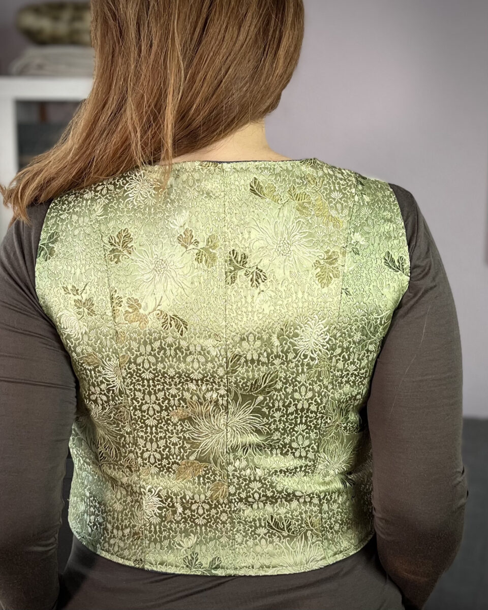 Back view of a white woman with red hair wearing a sage green silk brocade vest over an olive long sleeve t-shirt.