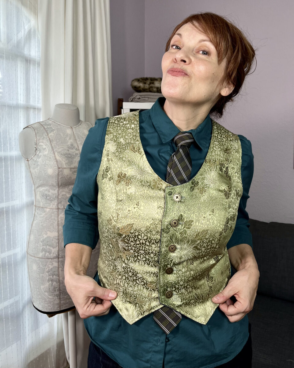 A white woman with red hair wears a sage green silk brocade vest over a teal button-down shirt with a grey, green, and white tie.