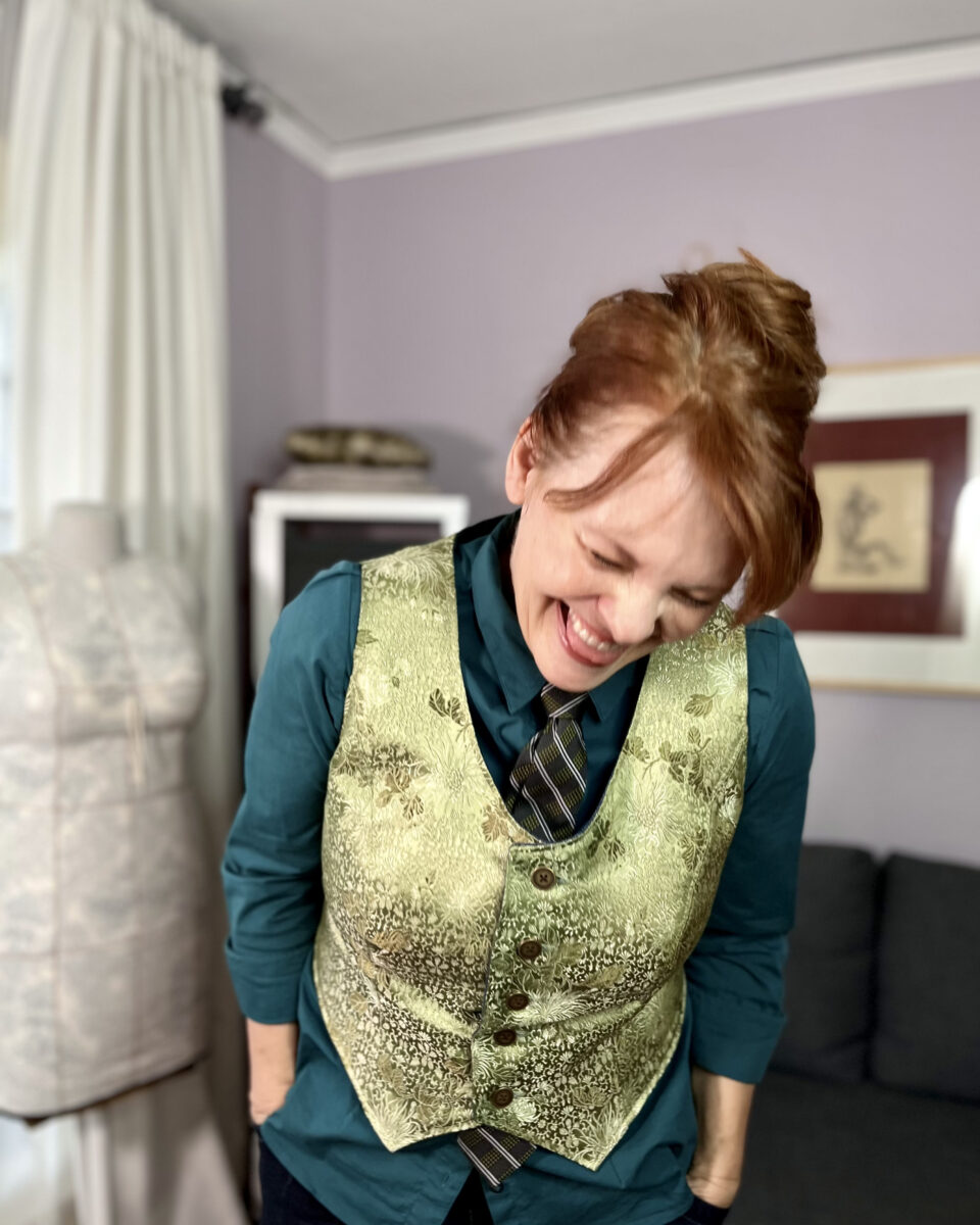 A laughing white woman with red hair wears a sage green silk brocade vest over a teal button-down shirt with a grey, green, and white tie.